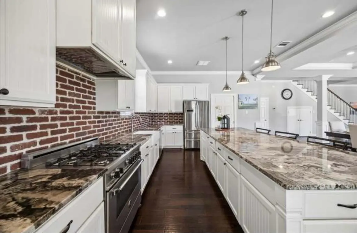 The guest kitchen with white cabinets, a brick backsplash, a gas range and a long stone island