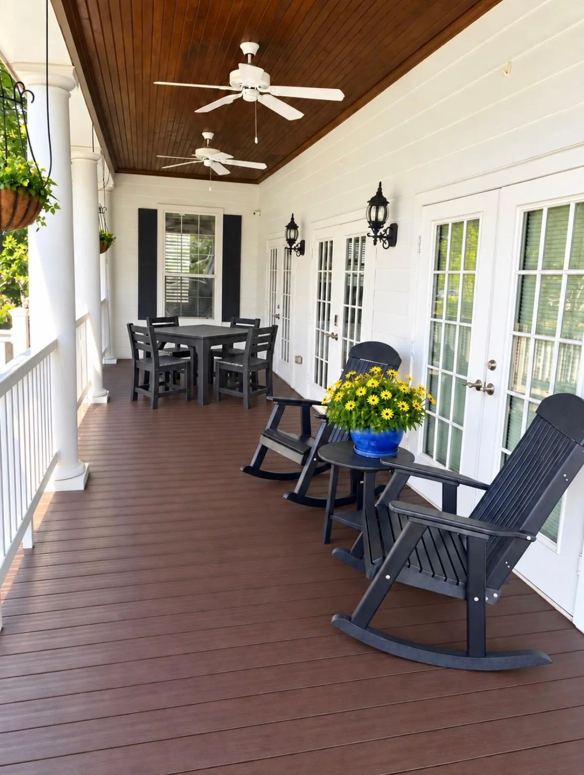 The covered porch of the guest home, with two black rocking chairs beside a pot of yellow flowers, a small dining table and chairs, ceiling fans and white columns